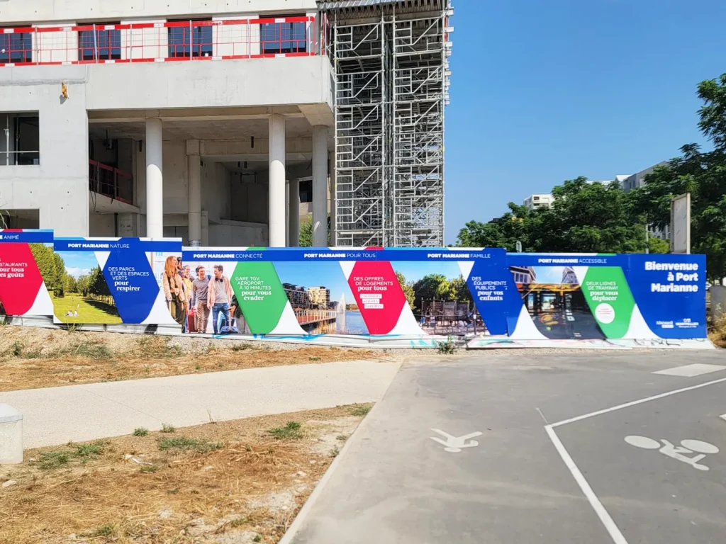 Habillage chantier à Port Marianne : palissade de chantier personnalisée aux couleurs du quartier devant un immeuble en construction à Montpellier